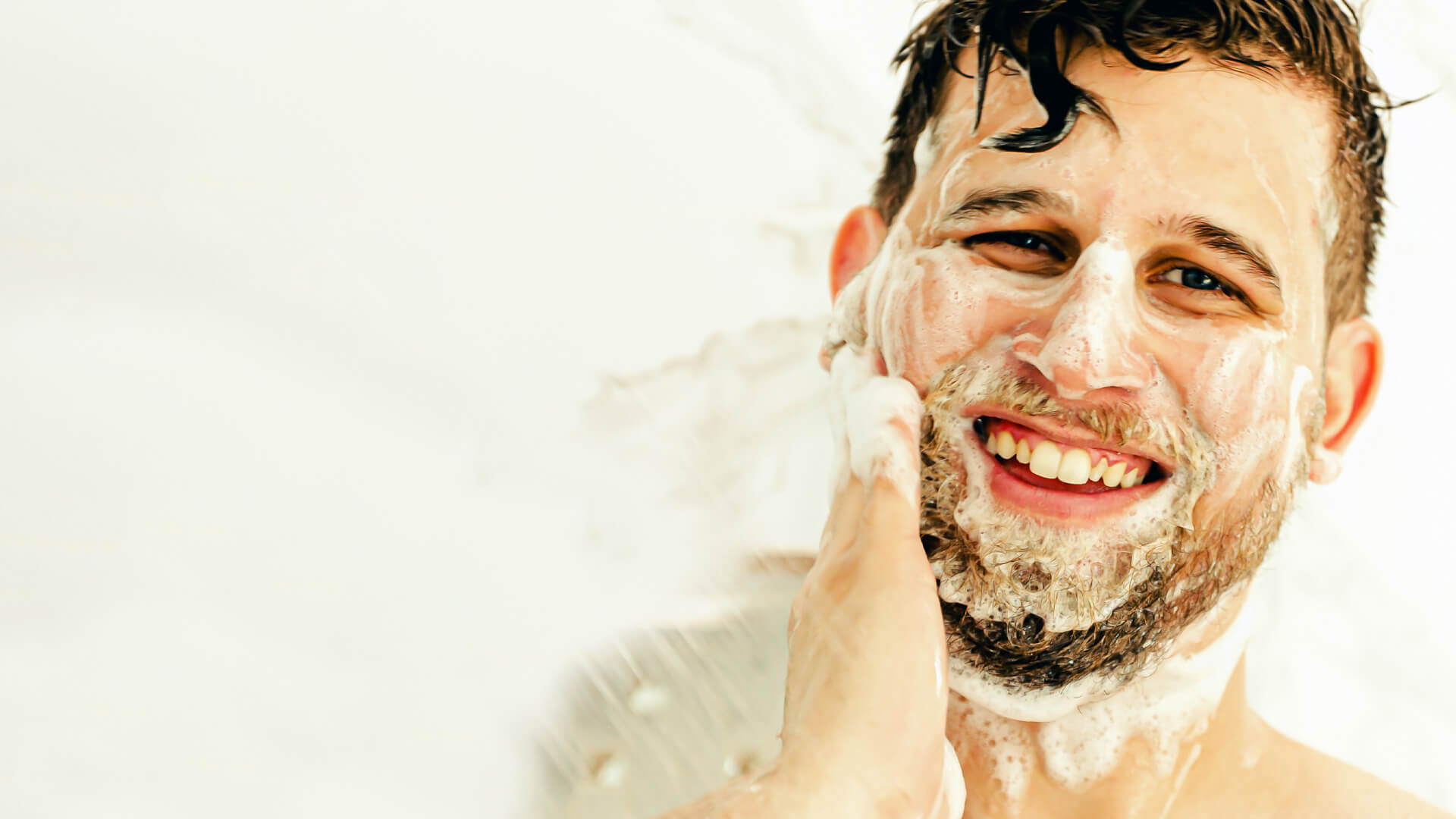 Close up of man in shower smiling while washing beard with suds